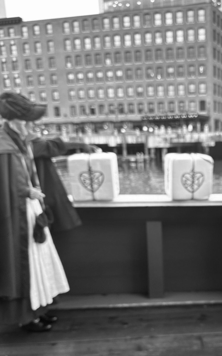 Period-costumed actor beside foam tea chest replicas on ship deck with harbor behind